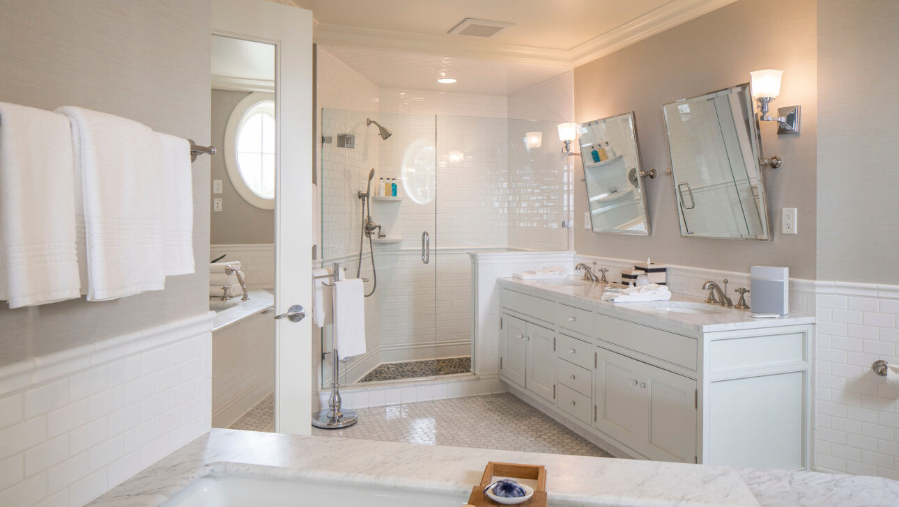 Bathroom with a white vanity, dual sinks, and a walk-in shower. Towels are neatly stacked, and a bathtub with a wooden tray is in the foreground.