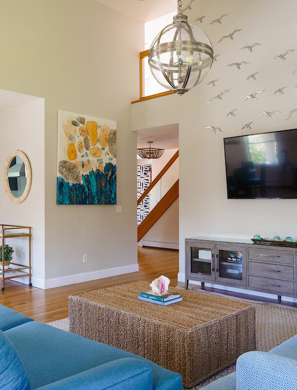 Modern living room with blue sofas, a large wall-mounted TV, and a decorative installation of birds on the wall. Adjacent kitchen with blue cabinets and wooden flooring.