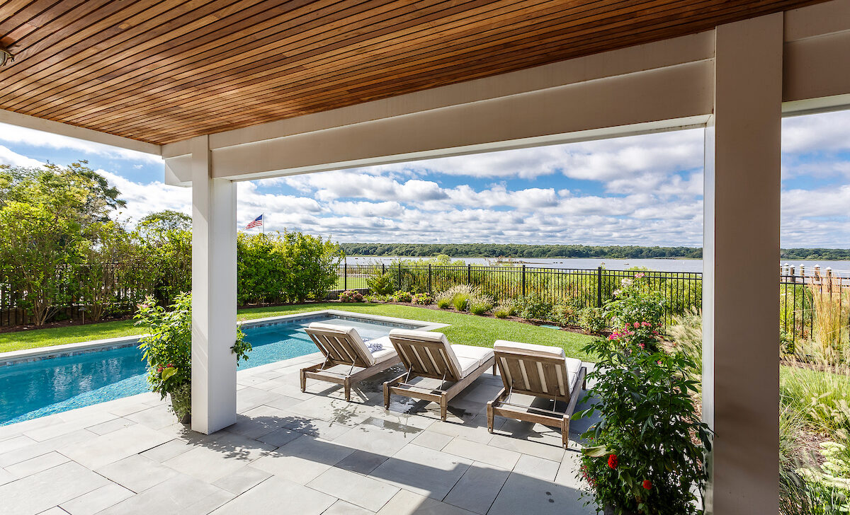 Patio with four wooden lounge chairs overlooks a pool and a scenic river view. Surrounded by greenery and clear skies.