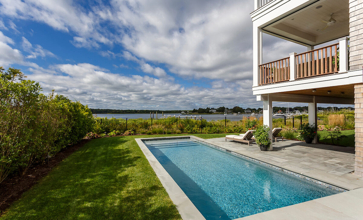 A backyard with a rectangular swimming pool, green lawn, and a house with a balcony overlooking a lake under a partly cloudy sky.