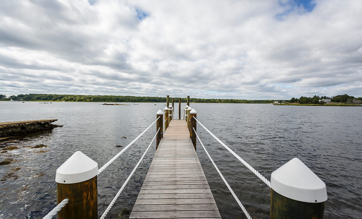 A wooden pier extends over a calm body of water under a cloudy sky, with green land visible in the distance.