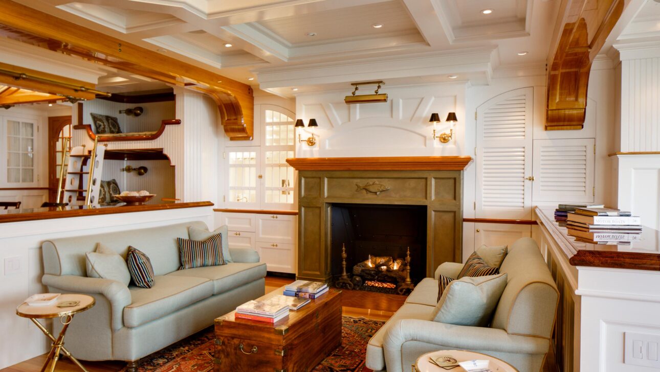 A cozy living room with two sofas facing a fireplace, wooden ceiling beams, and decorative shelves. A rug and small tables complete the seating area.