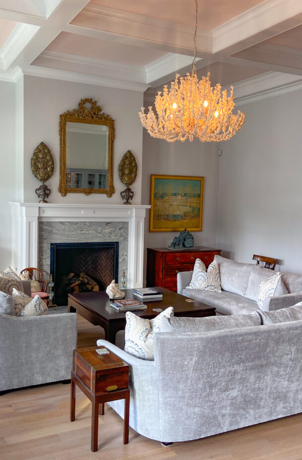 A bright living room with gray sofas arranged around a wooden coffee table. The room features a chandelier, decorative fireplace, and artwork on the walls.