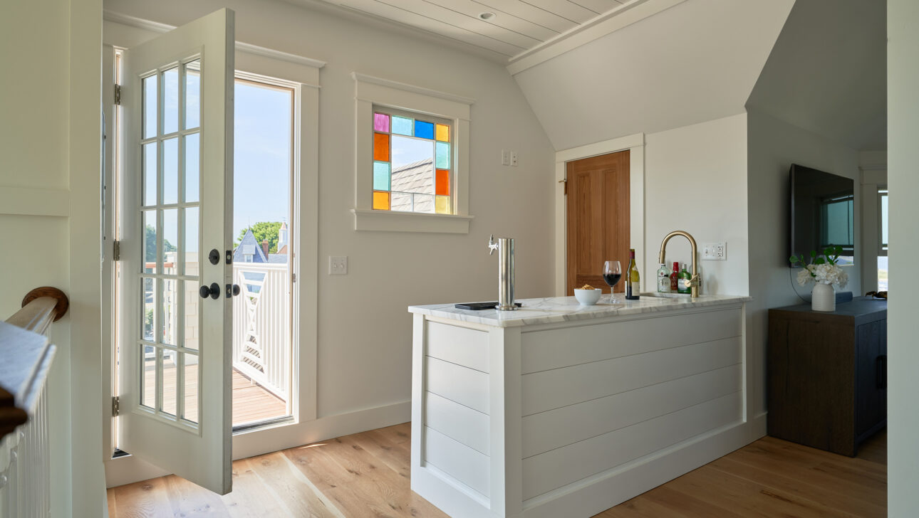 A small home bar with white paneling is by an open door leading to a deck. A glass of red wine and flowers are on the counter. A TV is visible in the corner.