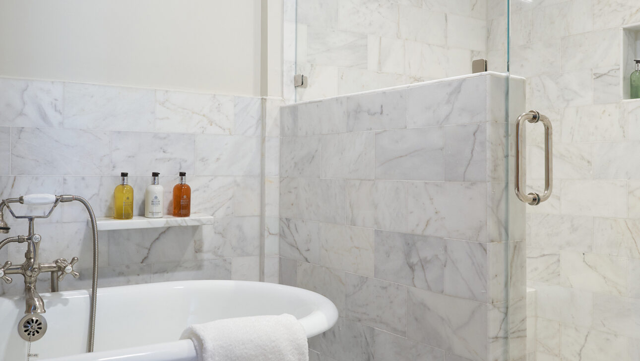 Marble bathroom with a freestanding tub, a towel draped over the side, and a glass-enclosed shower. Toiletries are on a small shelf.