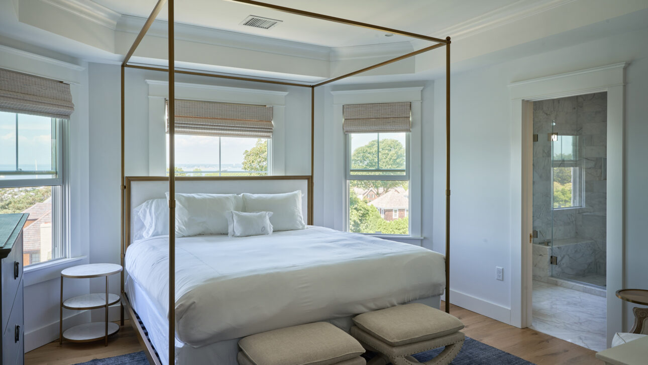 A bedroom with a canopy bed, white bedding, and two stools. The room has large windows with views of trees and water. A door on the right leads to a marble-floored bathroom.