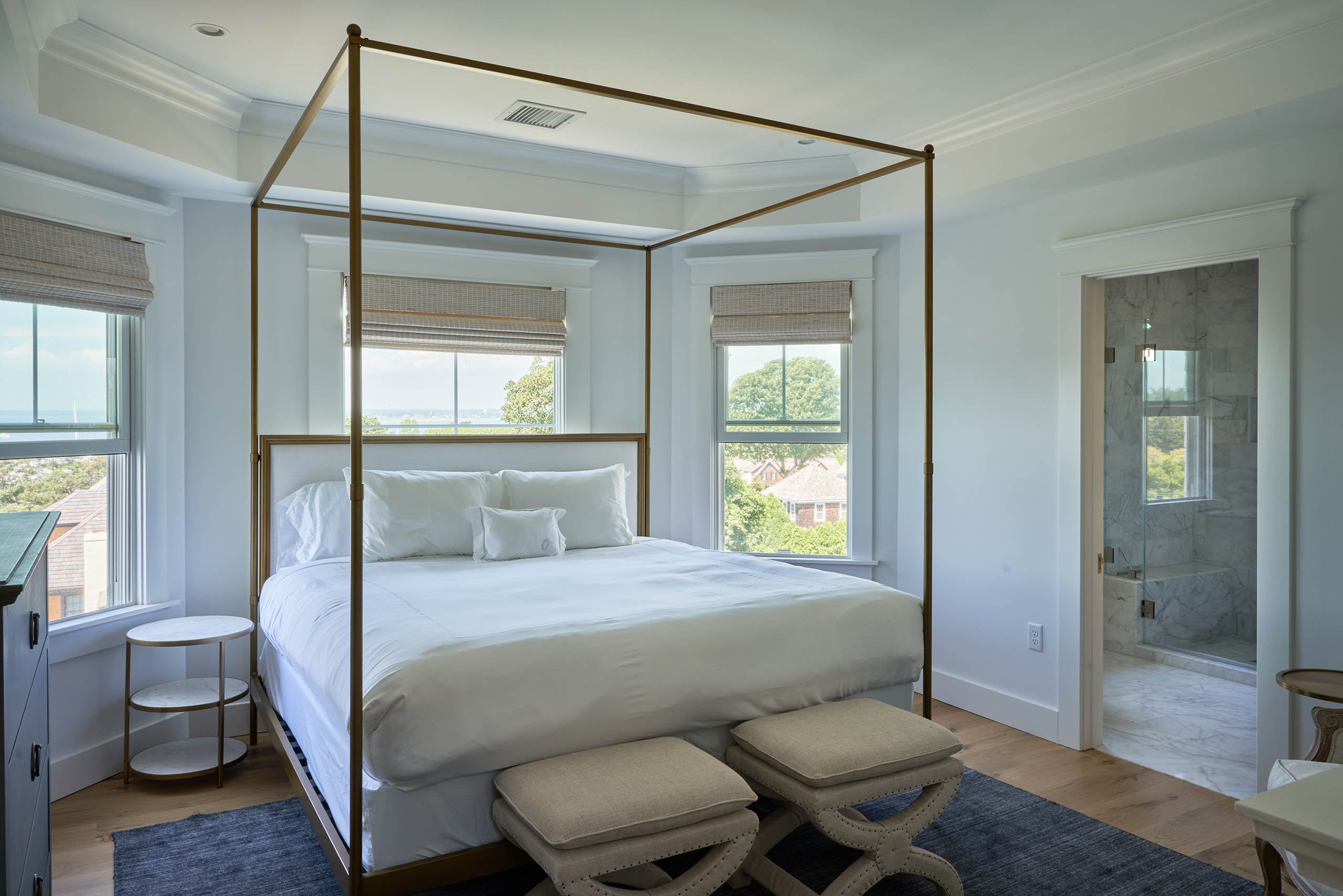 A bedroom with a canopy bed, white bedding, and two stools. The room has large windows with views of trees and water. A door on the right leads to a marble-floored bathroom.