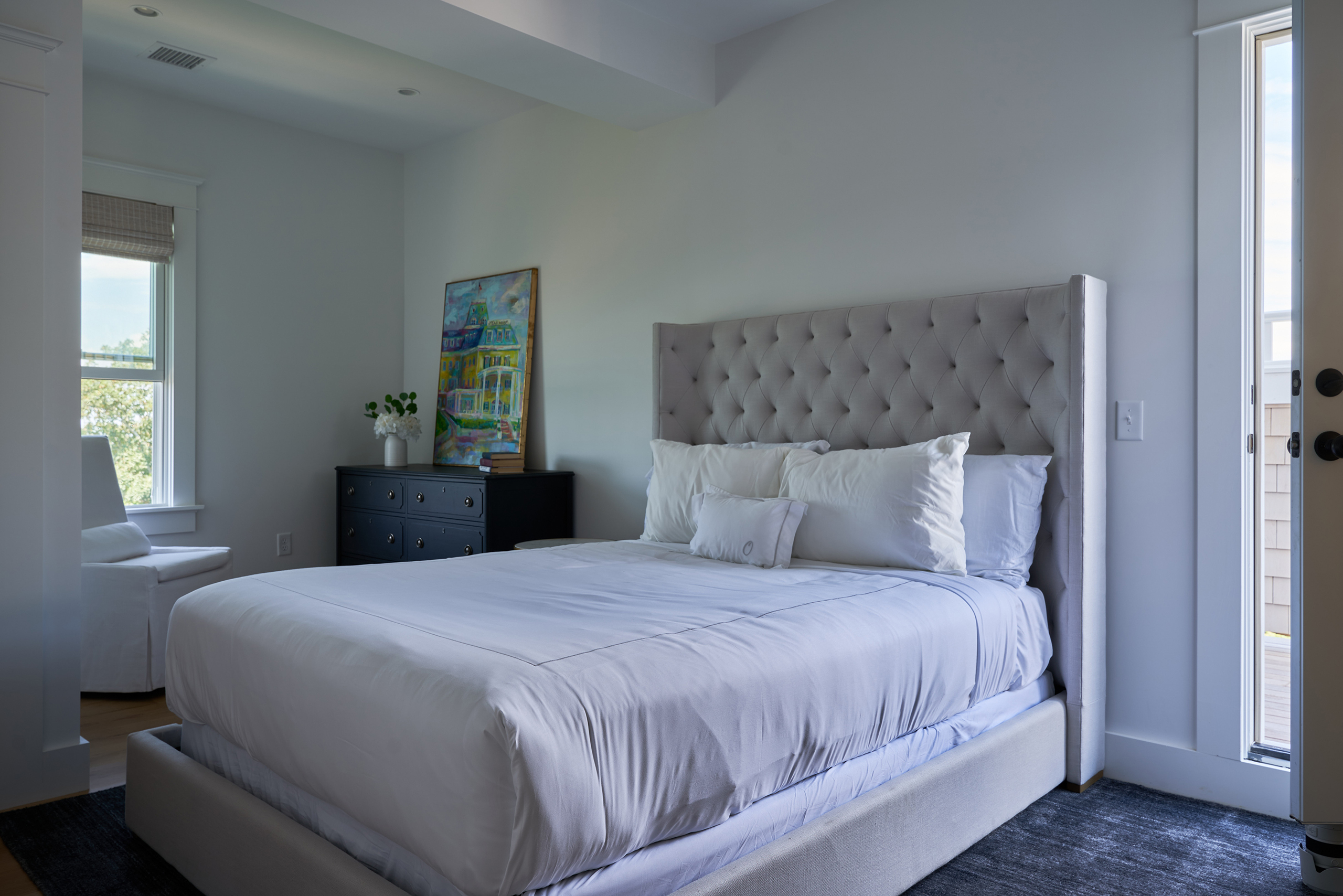 Bedroom with a neatly made bed, tufted headboard, white bedding, dresser, painting, and a window allowing natural light.