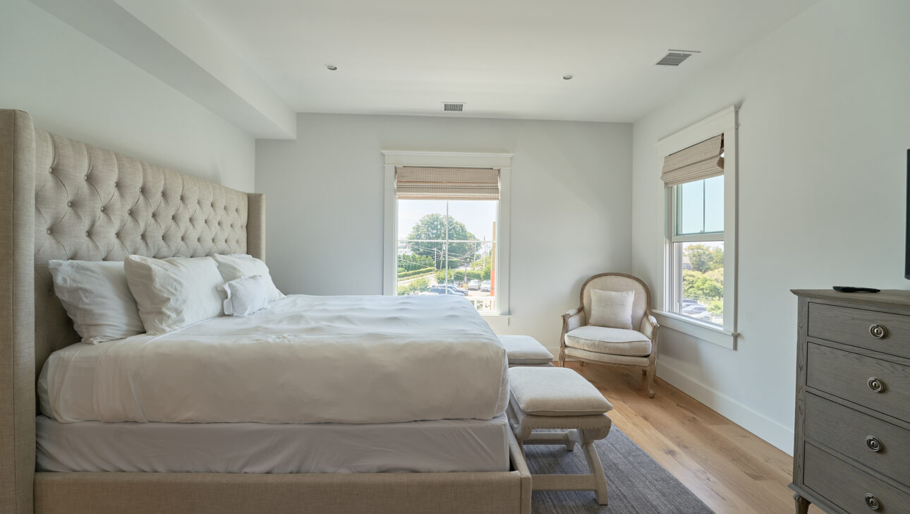 A bedroom with a large bed, light-colored bedding, tufted headboard, two windows with blinds, an armchair, and a dresser. The floor is wooden with a gray rug.