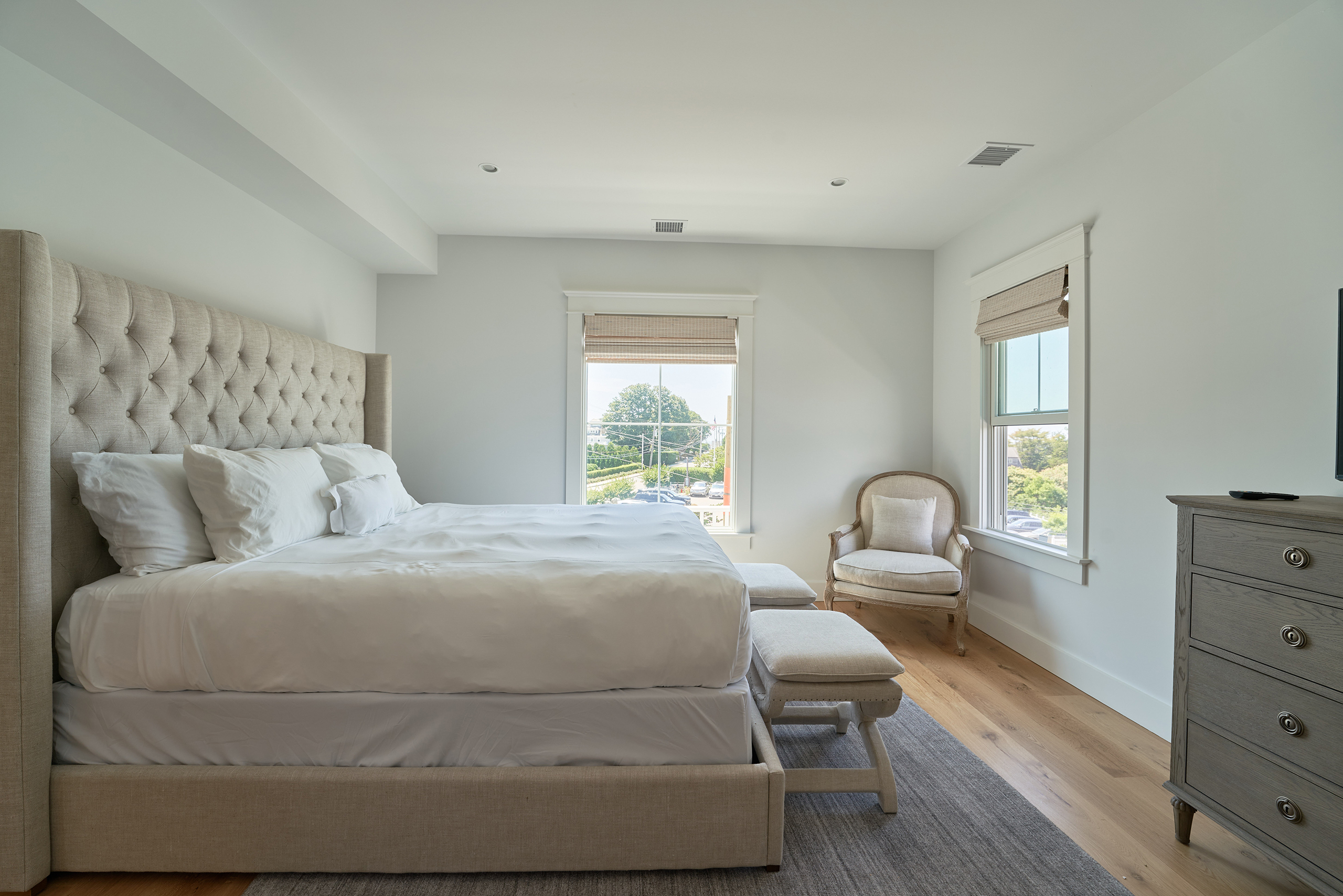 A bedroom with a large bed, light-colored bedding, tufted headboard, two windows with blinds, an armchair, and a dresser. The floor is wooden with a gray rug.