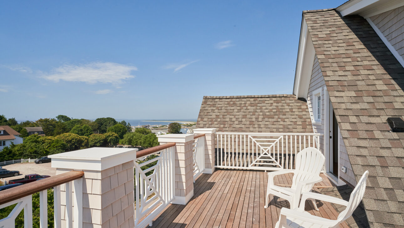 A rooftop deck with two white chairs overlooks a scenic view of trees and distant water under a clear blue sky.