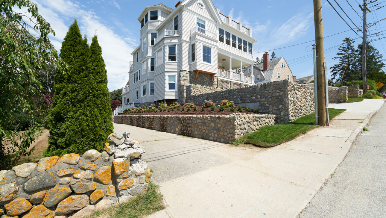 Large white house with multiple floors, surrounded by stone walls, on a sloped street under a clear blue sky.