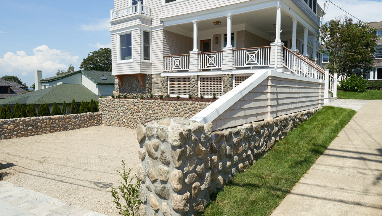 A large, two-story white house with a wraparound porch and stone wall, set against a blue sky.