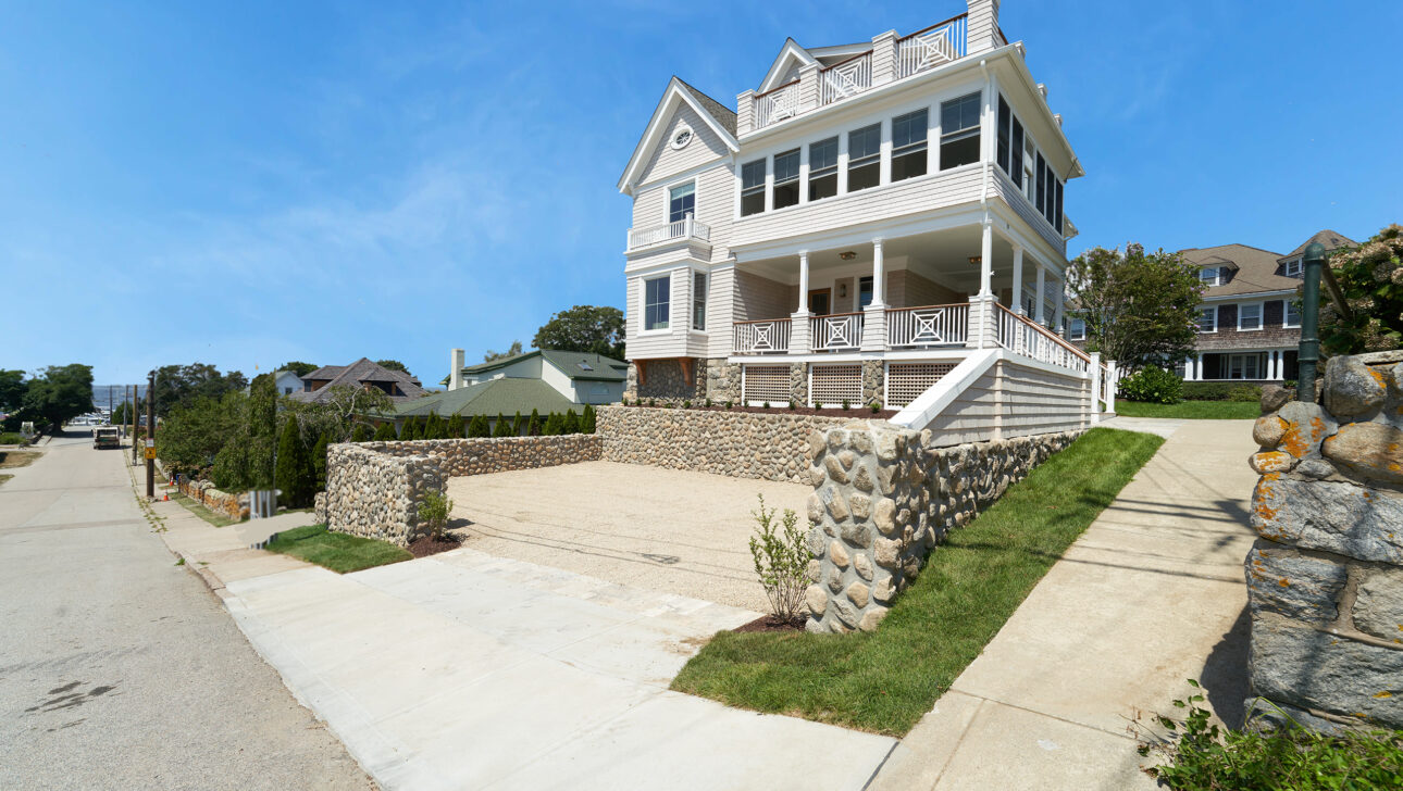 A large, white, three-story house with a stone retaining wall and driveway on a sunny day. The house has multiple gables and a wraparound porch.