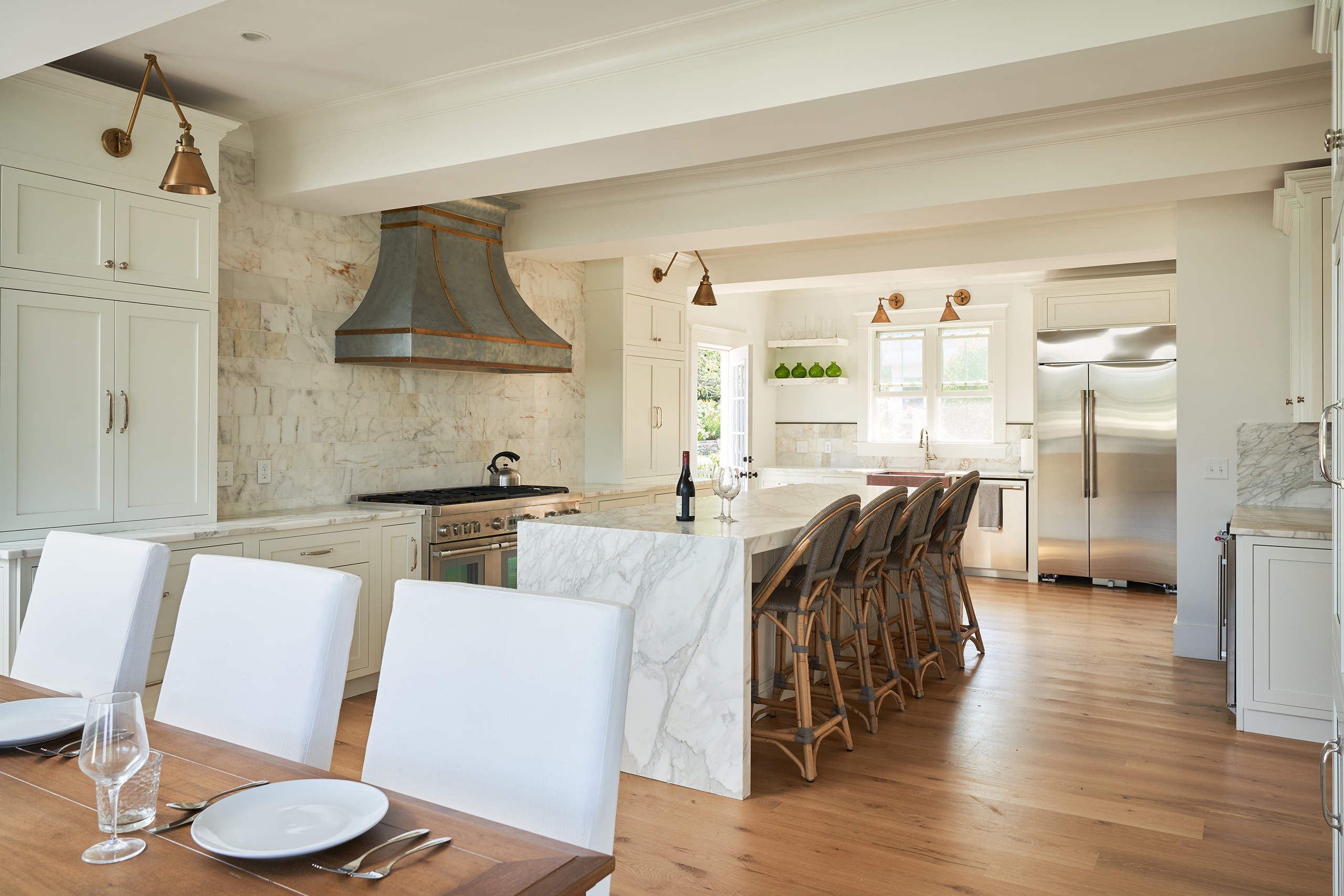 Spacious kitchen with a marble island, wicker bar stools, stainless steel appliances, and white cabinets. Wooden flooring and a table set with plates and glasses are visible.