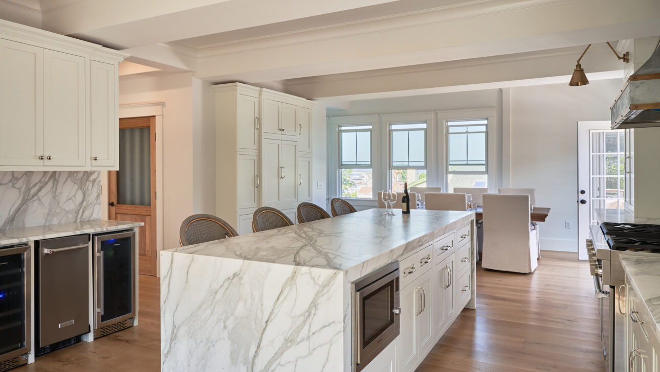 Modern kitchen with a marble island, bar stools, and white cabinetry. Wooden flooring and natural light fill the room. Dining area visible in the background.