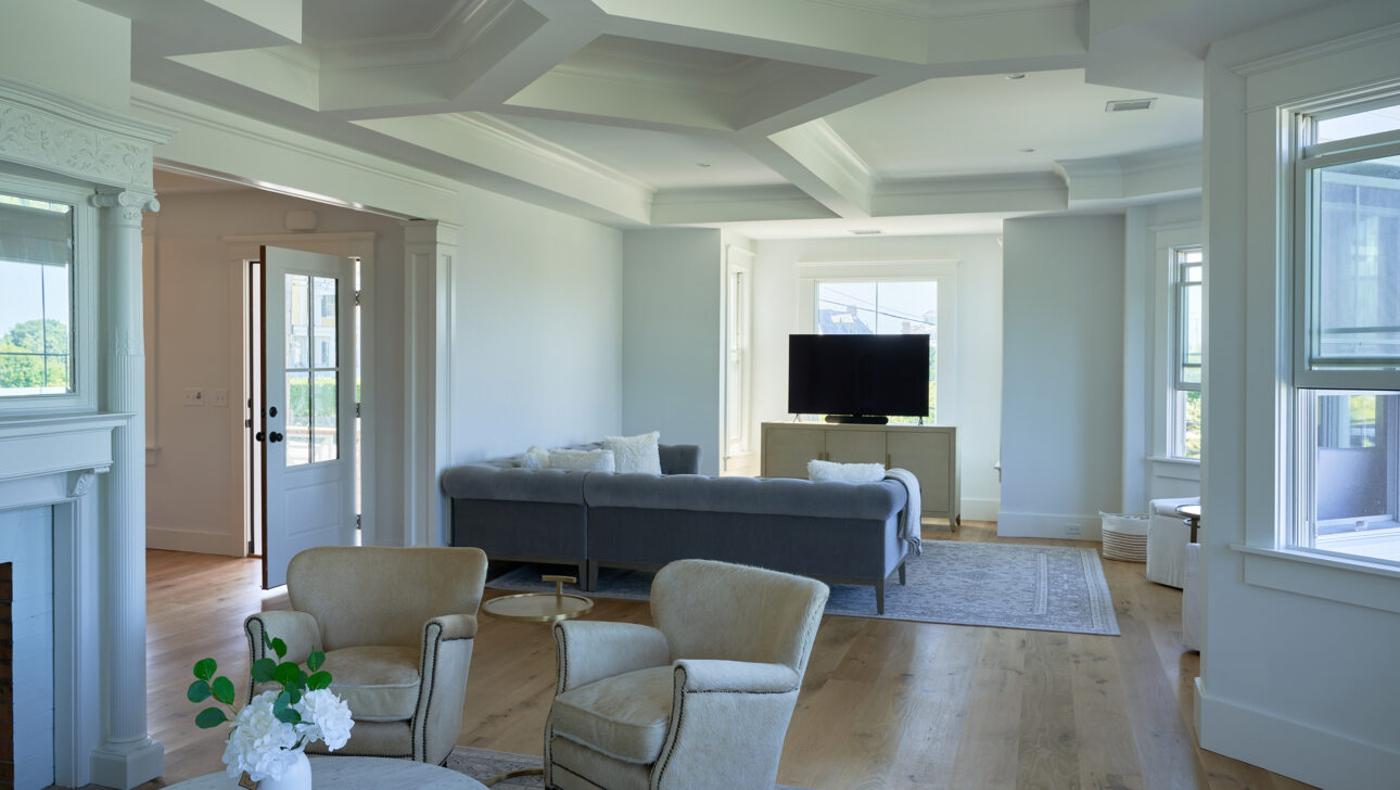 Spacious living room with light walls, wood flooring, and coffered ceiling. Features armchairs, a gray sofa, a wall-mounted TV, a round marble table with flowers, and large windows.