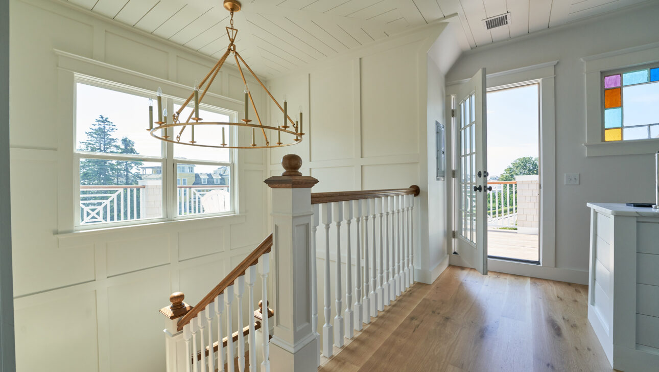 A bright staircase with white wooden railings leads to a hallway with large windows and an open door. A gold chandelier hangs from the ceiling.