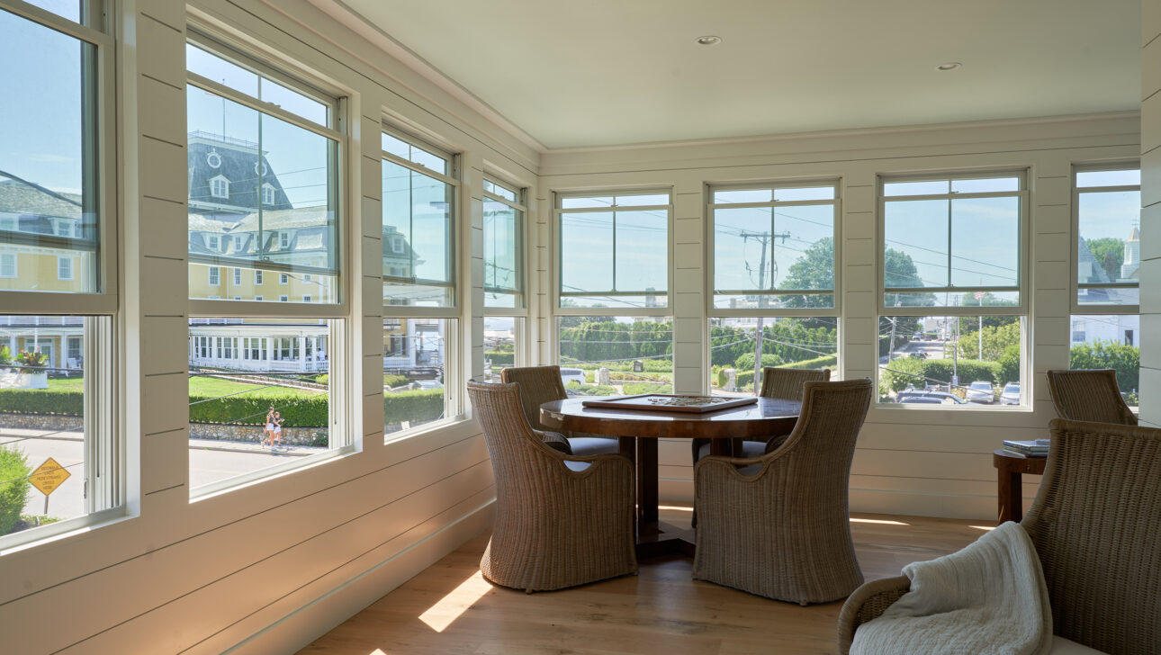 Sunlit room with wicker chairs around a wooden table, large windows offering views of a street and distant buildings, and light-colored wooden flooring.