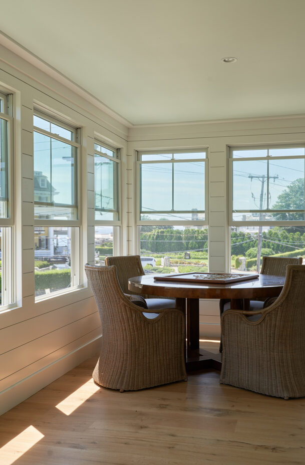Sunlit room with wicker chairs around a wooden table, large windows offering views of a street and distant buildings, and light-colored wooden flooring.