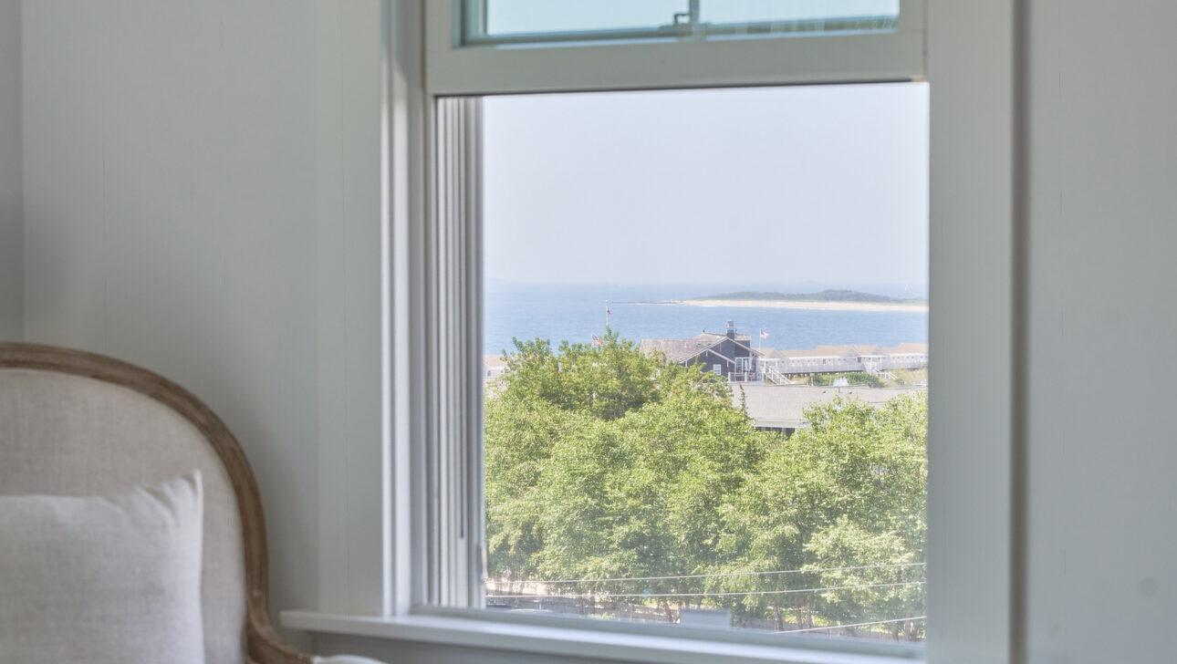 A window view from a bedroom shows a coastal scene with trees and the sea in the distance. An armchair and part of a bed are visible in the foreground.