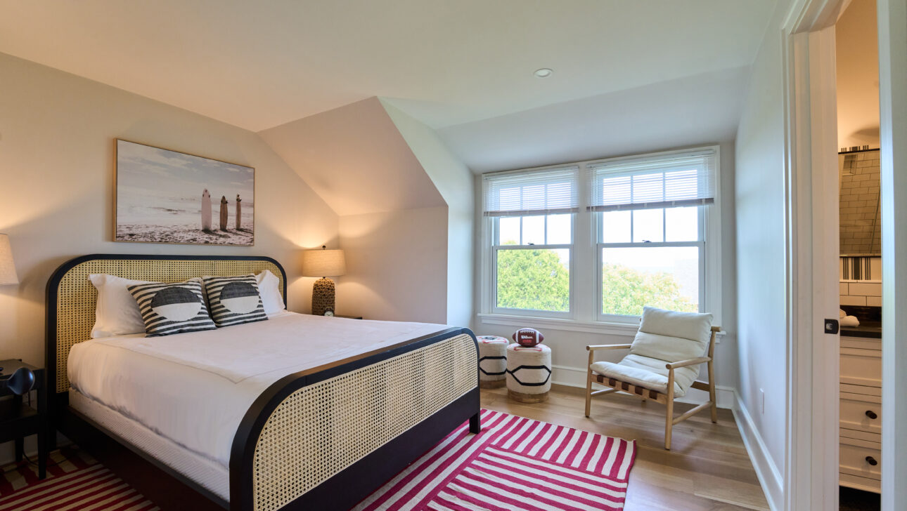 A bedroom with a double bed, striped pillows, a window with blinds, a chair, a painting above the bed, and two striped rugs on a wooden floor.