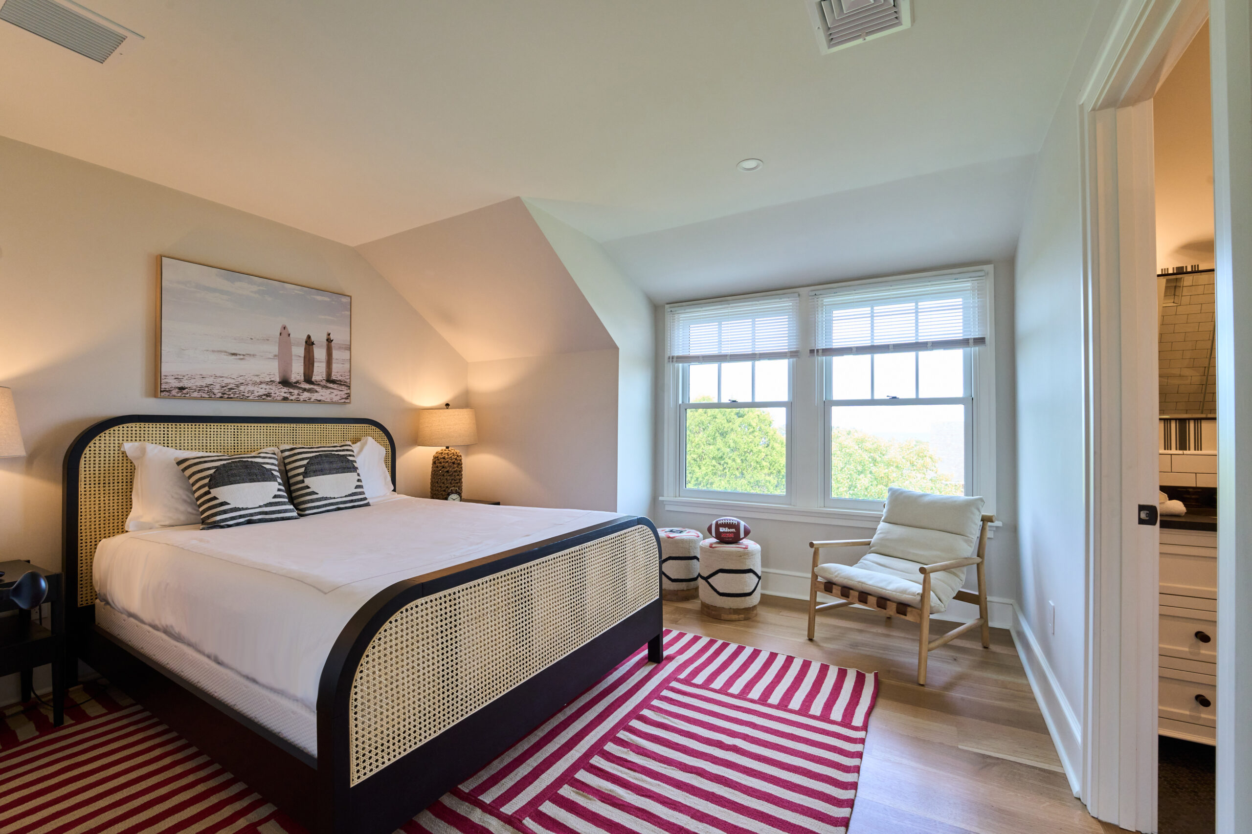 A bedroom with a double bed, striped pillows, a window with blinds, a chair, a painting above the bed, and two striped rugs on a wooden floor.