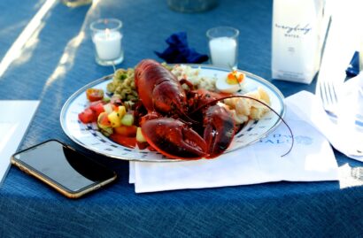 A cooked lobster on a plate with assorted sides, placed on a blue tablecloth beside a smartphone and a napkin.