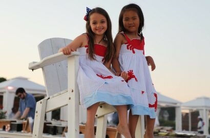 Two children in matching dresses sit on a white wooden lifeguard chair, smiling. The background features a man setting up tables and white tents.