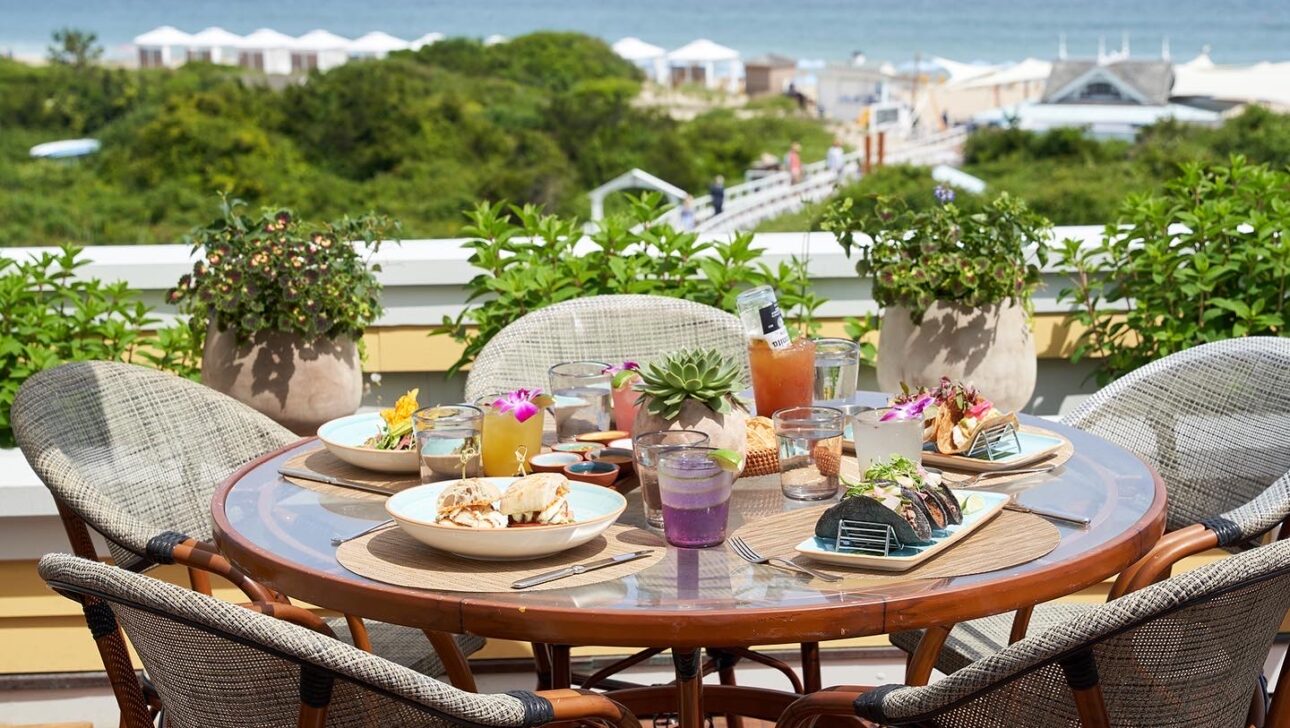 A round table with food and drinks overlooks a scenic coastal view with greenery, beach, and ocean in the background.