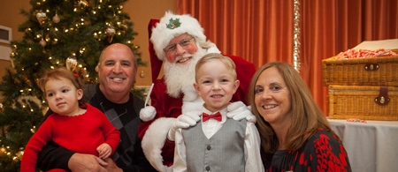 A family poses with Santa Claus in front of a Christmas tree. The child in the center wears a gray vest and red bow tie. The child on the left is in red. The adults are smiling.