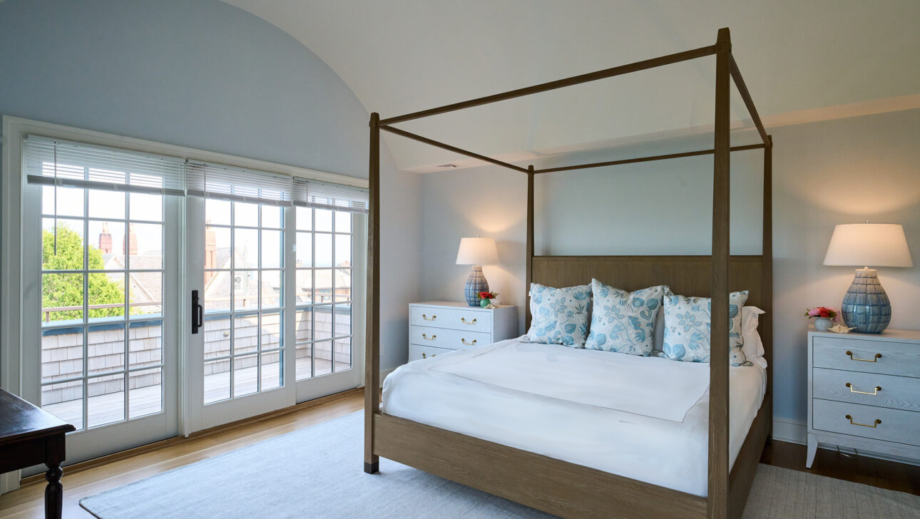 A spacious bedroom features a wooden four-poster bed with patterned pillows, flanked by two bedside tables with lamps, and a large window with glass doors leading to an outdoor area.