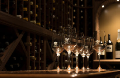 Five wine glasses sit on a wooden counter in a dimly lit wine cellar, with shelves of wine bottles in the background.