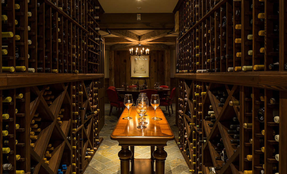 Dimly lit wine cellar with wooden shelves full of wine bottles, a wooden table with wine glasses, and a chandelier above.