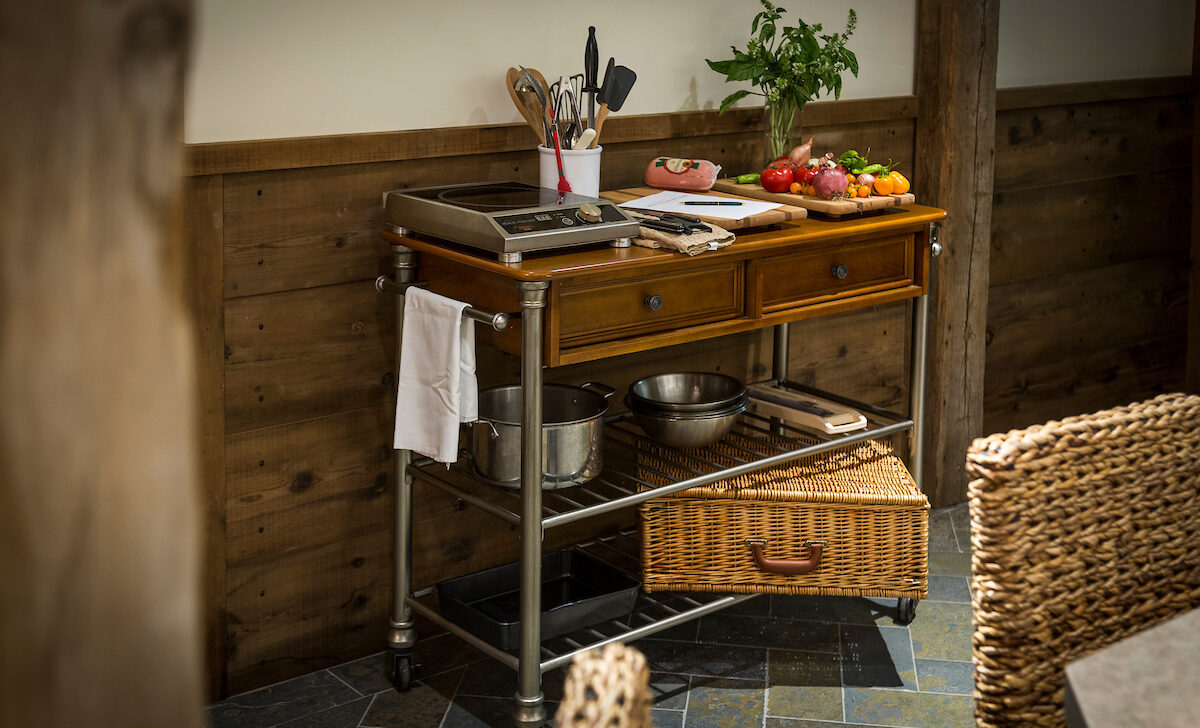 A wooden kitchen cart with a ceramic cooktop, utensils, vegetables, and baskets on a rustic tiled floor.