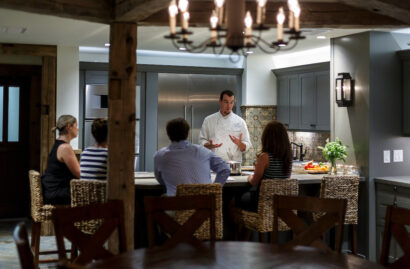 A chef in a white uniform giving a cooking demonstration to four seated people in a kitchen setting.