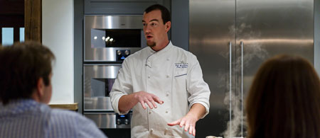 A chef in a white coat is speaking to two people in a kitchen. Steam rises from the cooking area.