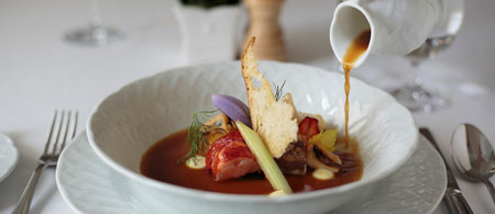A gourmet dish is plated in a white bowl with a variety of garnishes. A sauce is being poured from a small white pitcher into the bowl. The table is set with a fork, knife, and glass.