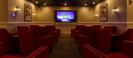 A small theater room features red chairs facing a screen displaying a movie scene. Movie posters adorn the walls, and soft lighting enhances the cozy atmosphere.