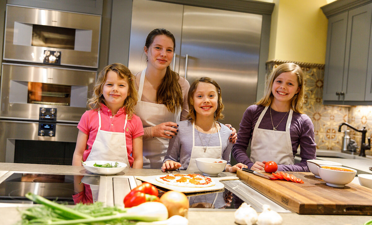 Four children in a kitchen, wearing aprons, are preparing food with vegetables on the counter.
