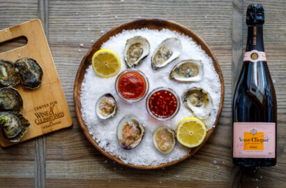 A platter of oysters on ice with lemon slices and dipping sauces sits beside a bottle of Veuve Clicquot Rosé champagne.