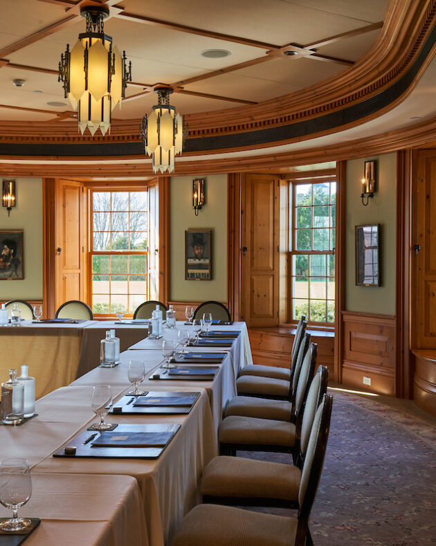 A conference room with a U-shaped table setup, chairs, and materials like notepads and water bottles. Large windows and ornate lighting fixtures are present. A blank flip chart stands in the corner.