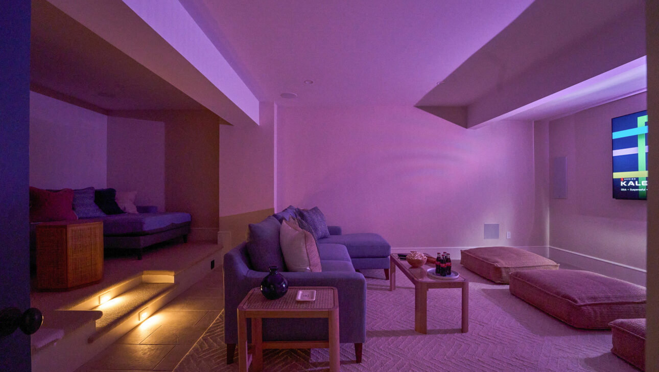 A dimly lit room with a sectional sofa, floor cushions, stairs with step lights, and a wall-mounted TV displaying a paused screen. Tables hold decor items.