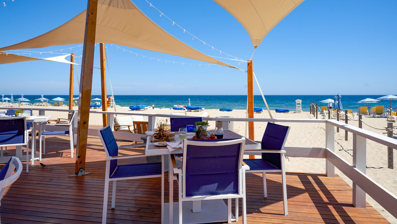 Beachside dining area with purple chairs and wooden tables under sail shades, overlooking a sandy beach and the ocean. Umbrellas and lounge chairs are visible in the background.