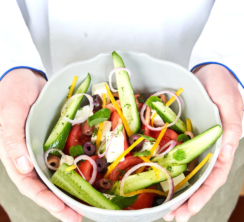 Person holding a bowl of fresh salad with cucumber, olives, red onions, cheese, peppers, and herbs.