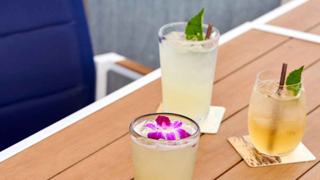 Three cocktails on a wooden table, each in a glass with a coaster. One drink has an orchid garnish, another with a mint leaf. A blue cushioned chair is visible on the left.