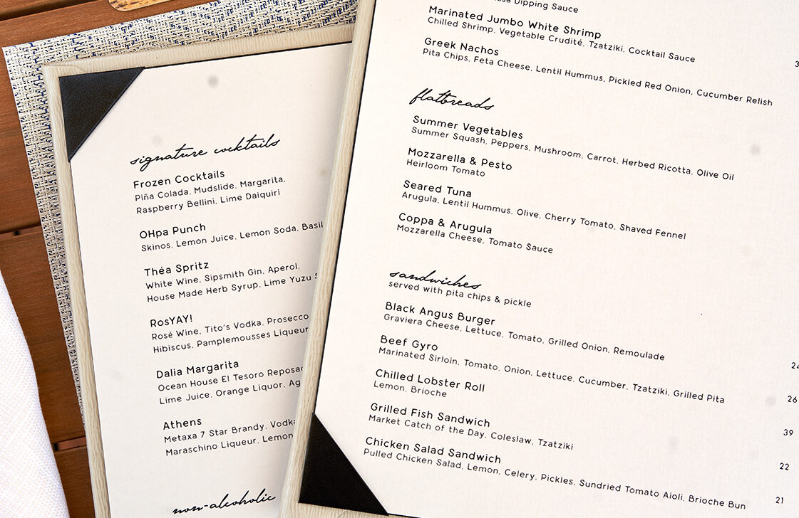 A menu on a wooden table next to a drink with a purple flower. The menu lists various cocktails, salads, and entrees.
