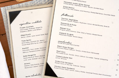 A menu on a wooden table next to a drink with a purple flower. The menu lists various cocktails, salads, and entrees.