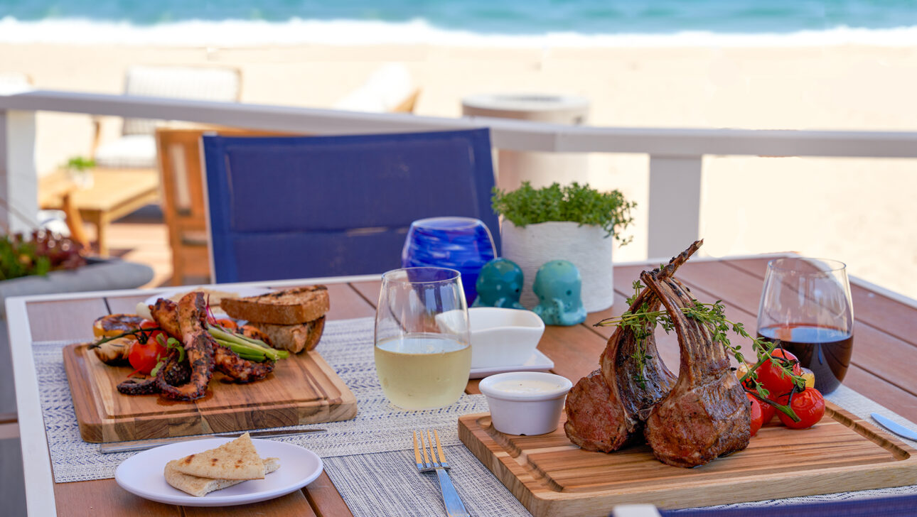 Outdoor dining setup with grilled lamb chops, vegetables, bread, and sauces on wooden boards. Two glasses of wine are on the table, with a beach and ocean visible in the background.