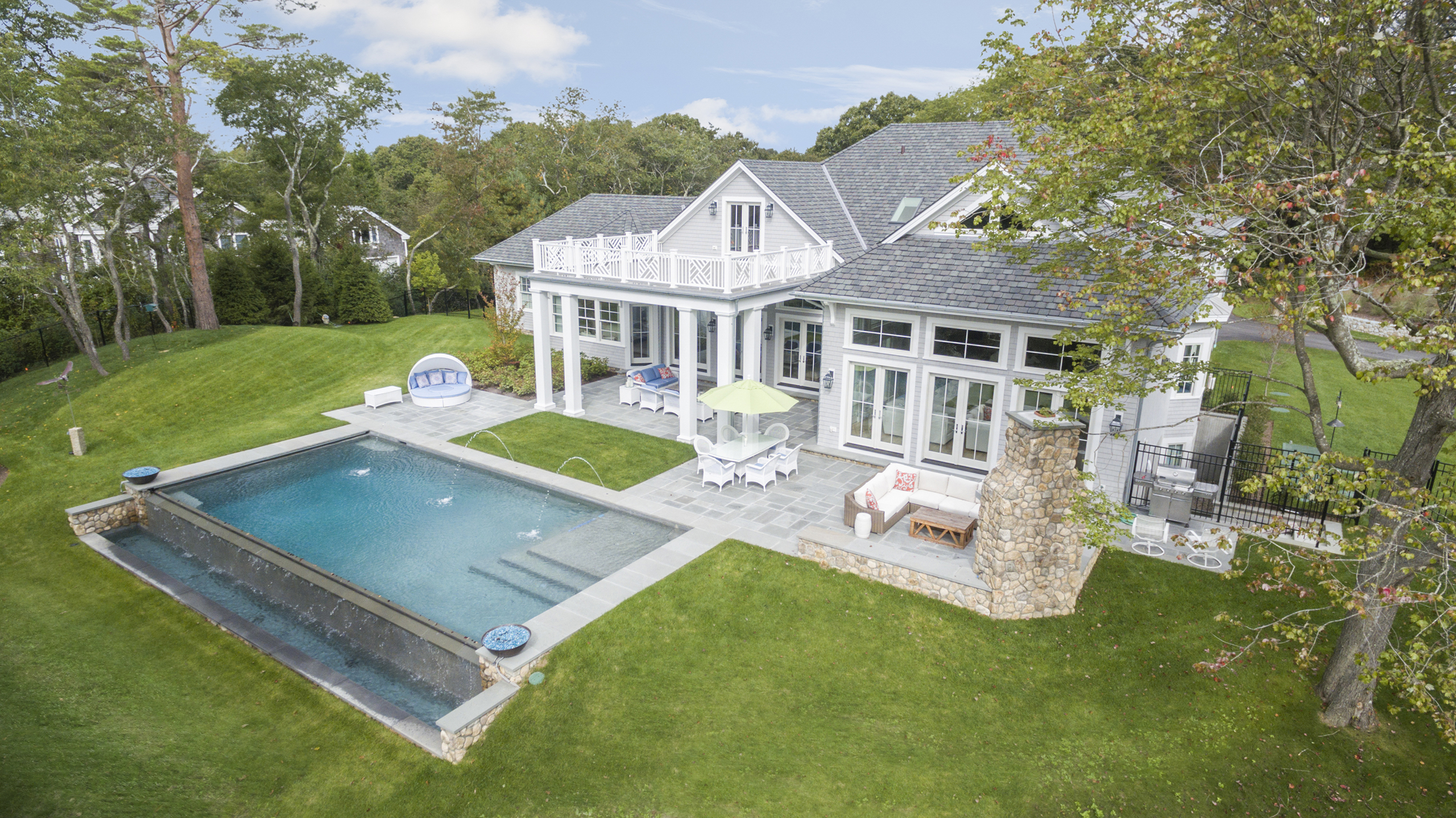 Aerial view of a large, modern house with a spacious patio, outdoor seating, and a rectangular infinity pool surrounded by well-manicured lawns and trees.
